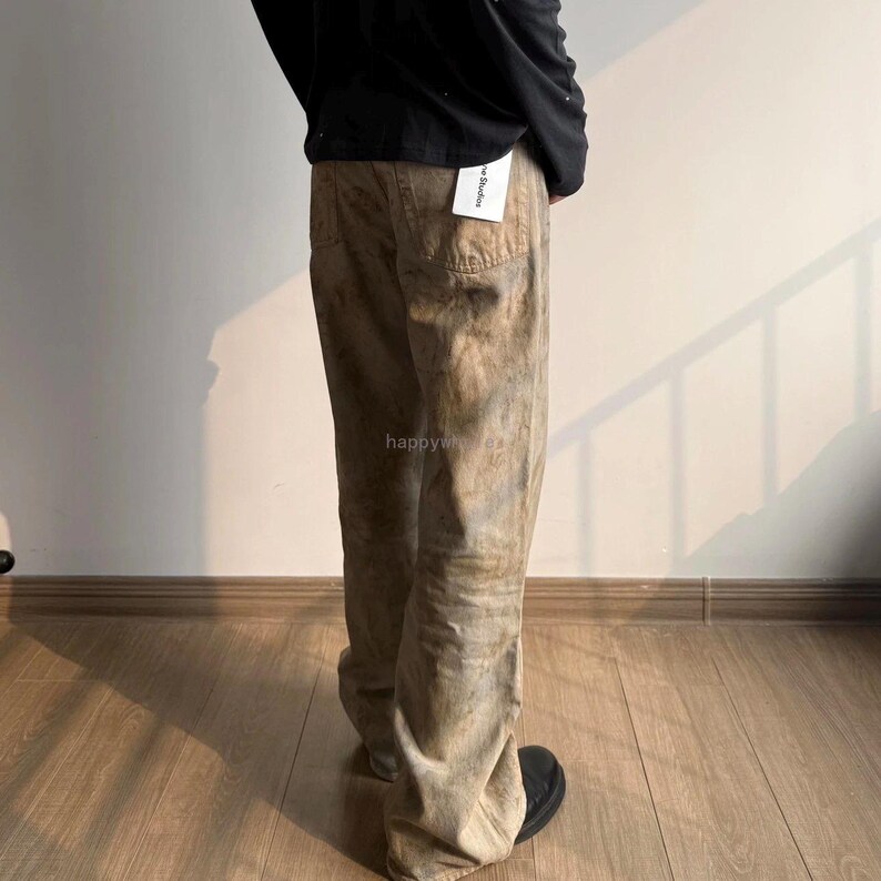 May include: A pair of tan and brown distressed denim jeans with a mottled, washed-out appearance. The jeans feature a straight-leg cut, a back pocket, and a white brand tag attached to the waistband. A black long-sleeve shirt is worn above.