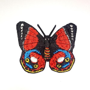 Puede incluir: Un parche bordado de mariposa con base negra y detalles en hilos rojo, azul y amarillo. Las alas muestran patrones simétricos con acentos blancos, mientras que el cuerpo central presenta rayas con costuras en rojo y amarillo.