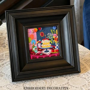 以下が含まれることがあります： ケーキ、風船、花のある誕生日のシーンを描いた額入りの刺繍。刺繍は正方形の黒いフレームに入っています。テキスト「EMBROIDERY DECORATIVE」はフレームの下部にあります。