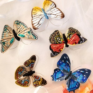 Könnte beinhalten: Fünf gestickte Schmetterlinge in verschiedenen Farben und Mustern, darunter Blau, Rot, Gelb und Braun, auf weißem Hintergrund.