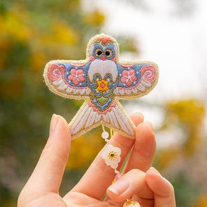 May include: A colorful embroidered owl kite with pink flowers and gold accents. The kite is hanging from a string with a white flower charm.