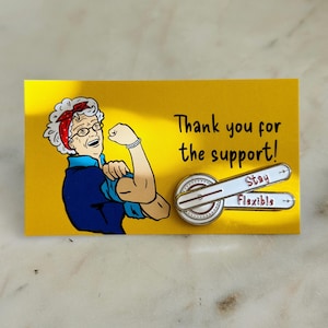 May include: A yellow card with a cartoon illustration of a woman flexing her bicep. The card says "Thank you for the support!" and features a white enamel pin with a red and white dial and the words "Stay Flexible".