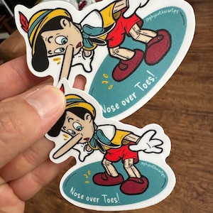 Pode incluir: Dois adesivos com um Pinóquio de desenho animado com um nariz comprido, vestindo uma roupa vermelha e amarela, e pernas de madeira. Os adesivos estão sobre um fundo azul com o texto "Nose over Toes!" impresso neles.