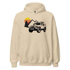 May include: A beige hoodie featuring a black graphic of a 4x4 vehicle, mountains, and a sunset. The hoodie has a hood with a drawstring and a kangaroo pocket in the front. The graphic is on the front of the hoodie.
