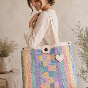May include: A large rectangular tote bag featuring a patchwork design of vertical stripes and small squares in pastel yellow, pink, blue, purple, and green floral patterns. The bag has a cream-colored top border, a dark button closure, and a heart charm.