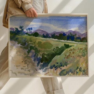 May include: Watercolor landscape painting in a light wooden frame. The artwork depicts a vibrant scene with rolling green hills, purple mountains, and a blue sky. The painting has a soft, natural aesthetic.