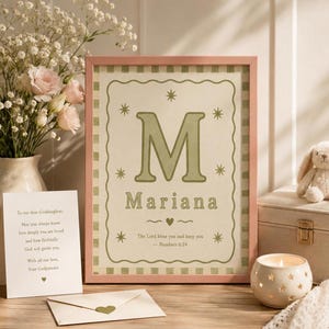 May include: A framed print with the name "Mariana" and the letter "M" in green, surrounded by stars. A vase of flowers, a card, an envelope, a candle, and a stuffed bunny are on a wooden surface.