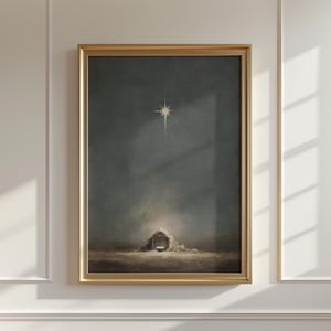 May include: A framed print of a nativity scene. The artwork shows a star shining above a small structure, with a muted colour palette of dark blues and browns. The frame is gold.
