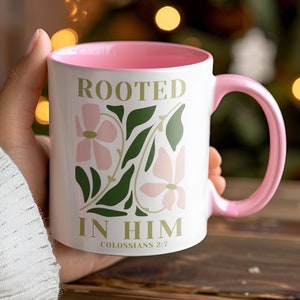 Op de afbeelding: Een roze en witte keramische mok met een bloemenontwerp en de tekst "Rooted in Him" en "Colossians 2:7".