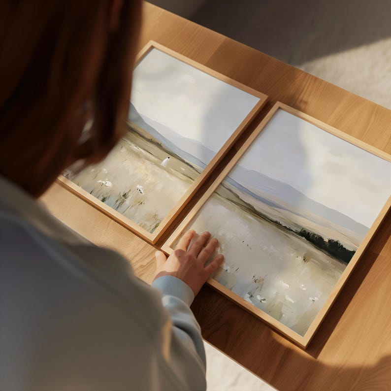 May include: Two framed landscape art prints. Each print features a serene field scene with soft colors, a light blue sky, and distant mountains. The frames are made of light-colored wood, and the overall aesthetic is minimalist and calming.