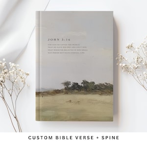 May include: A hardcover book with a light gray and olive green landscape painting on the cover. The text John 3:16 is printed in black, followed by the verse: For God so loved the world that he gave his one and only son, that whoever believes in him shall not perish but have eternal life.
