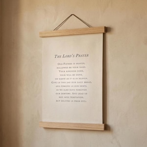 May include: A canvas print of "The Lord's Prayer" in black text, framed by natural wood bars and suspended by a cord. The background is a neutral beige. The print is a simple, elegant piece of wall art.