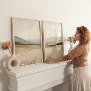 May include: Two framed landscape art prints with neutral tones of beige, green, and white. The artwork depicts a field with a figure and mountains in the background. The frames are light wood, and the prints are displayed on a white mantel.