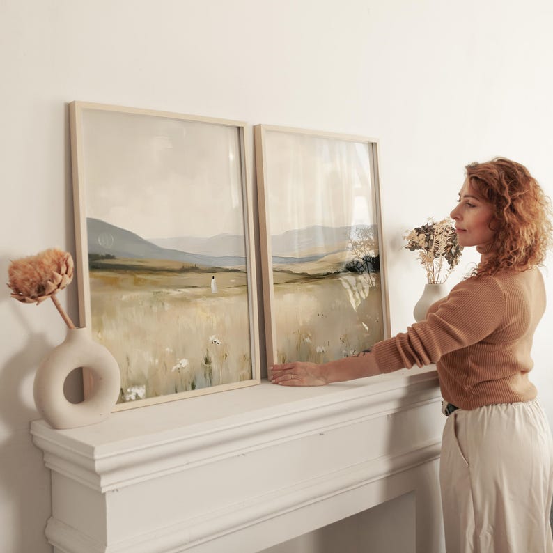 May include: Two framed landscape art prints with neutral tones of beige, green, and white. The artwork depicts a field with a figure and mountains in the background. The frames are light wood, and the prints are displayed on a white mantel.