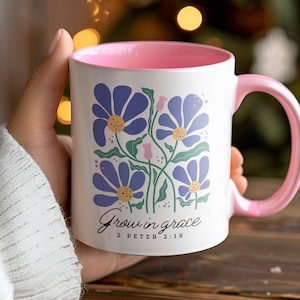 Op de afbeelding: Witte keramische mok met een roze rand en handvat. De mok heeft een bloemenmotief met blauwe bloemen en groene bladeren. De tekst "Grow in grace 2 Peter 2:18" staat op de mok gedrukt.