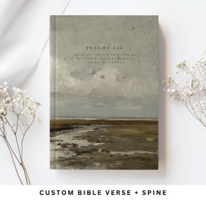 Könnte beinhalten: Ein Hardcover-Buch mit einem Landschaftsgemälde auf dem Einband. Das Kunstwerk zeigt einen bewölkten Himmel über einer Küstenlinie in gedämpften Tönen von Grau, Braun und Beige. Der Text auf dem Einband lautet "PSALMS 144" und ein Vers. Der untere Teil des Buches lautet "CUSTOM BIBLE VERSE + SPINE."