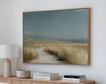 Könnte beinhalten: Ein gerahmtes Landschaftsgemälde zeigt eine Person, die auf einem Pfad durch hohes, goldenes Gras auf ein Gewässer unter einem bewölkten Himmel zugeht. Das Kunstwerk befindet sich in einem hellen Holzrahmen, der auf einem hellen Holzschrank montiert ist. Bücher und eine Lampe sind ebenfalls sichtbar.
