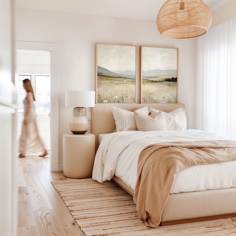 May include: A bedroom with a neutral color palette. A bed with a beige headboard, white bedding, and a tan throw blanket. Two landscape paintings hang above the bed. A woven pendant light hangs from the ceiling. A woman walks through a doorway.