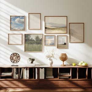 May include: A gallery wall featuring framed art prints with landscape and text designs. A dark wood cabinet below displays books, decorative objects, and a woven lamp. The color palette includes natural tones and earth colors.