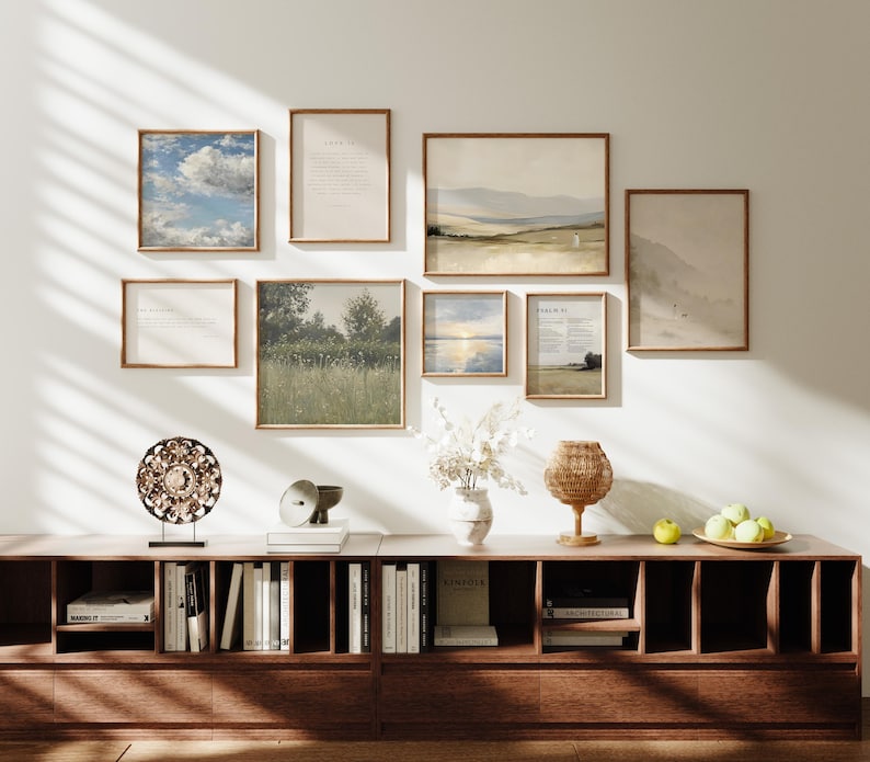 May include: A gallery wall featuring framed art prints with landscape and text designs. A dark wood cabinet below displays books, decorative objects, and a woven lamp. The color palette includes natural tones and earth colors.