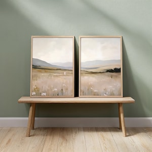 May include: Two framed landscape paintings with a neutral color palette. The artwork depicts a field with mountains in the background. The frames are made of light-colored wood. The paintings are displayed on a wooden bench.
