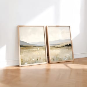 May include: Two framed landscape art prints. The artwork depicts a field of tall grass with a figure in the distance, mountains, and a cloudy sky. The frames are light wood, and the overall color palette is neutral.