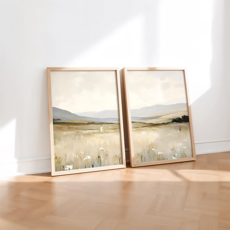 May include: Two framed landscape art prints. The artwork depicts a field of tall grass with a figure in the distance, mountains, and a cloudy sky. The frames are light wood, and the overall color palette is neutral.