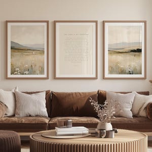 Puede incluir: Tres impresiones artísticas enmarcadas con paisajes en tonos neutros. Las impresiones presentan campos, montañas y una cita del Salmo 23. Los marcos son de color marrón claro. La obra de arte se exhibe sobre un sofá marrón con cojines decorativos.