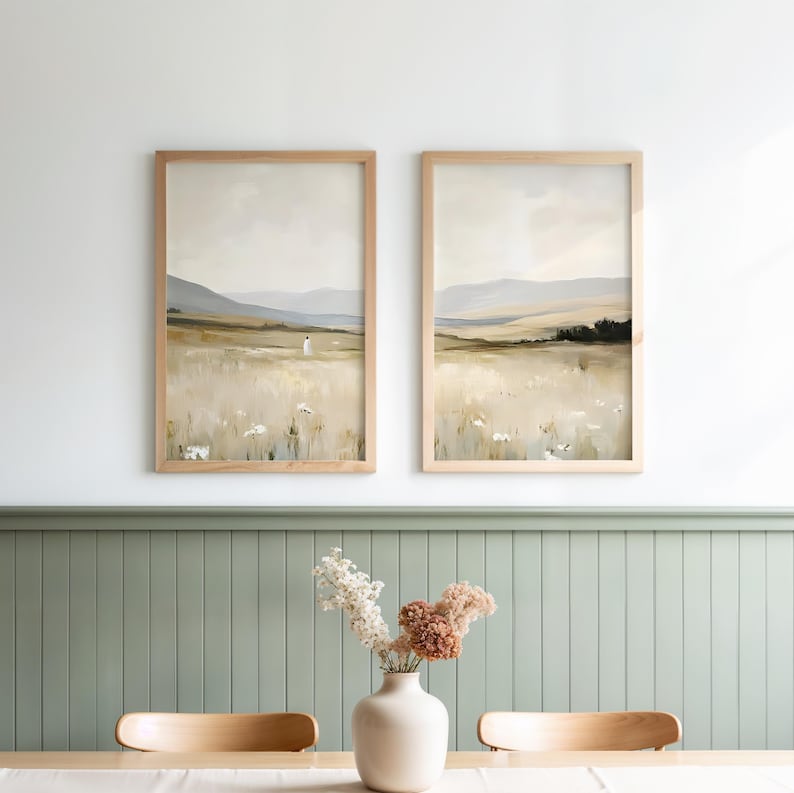 May include: Two framed landscape paintings with light wood frames. The artwork depicts a field with mountains in the background. A vase with dried flowers sits on a table in front of the paintings. The color palette is neutral with beige, brown, and green tones.