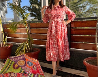 May include: A red and white patterned dress with a collared neckline and a matching belt. The dress is knee-length and has three-quarter sleeves. A colorful, patterned umbrella is on the ground. The person is wearing sandals.