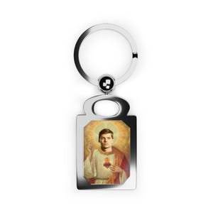 May include: A silver keychain with a rectangular charm featuring a man with a heart in his hand. The man is wearing a white robe with a red sash and a gold halo. The background is a yellow and orange gradient.
