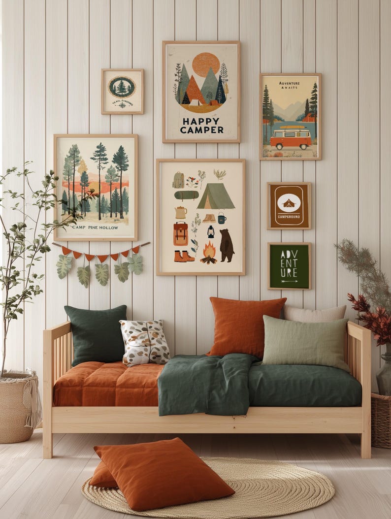 Puede incluir: Una escena de dormitorio acogedora con un sof&aacute; cama de madera con ropa de cama y cojines naranjas y verdes. Encima, arte enmarcado con tem&aacute;tica de camping con textos como "Happy Camper" y "Adventure Awaits" decora la pared. Una alfombra tejida y plantas en macetas completan el espacio acogedor.