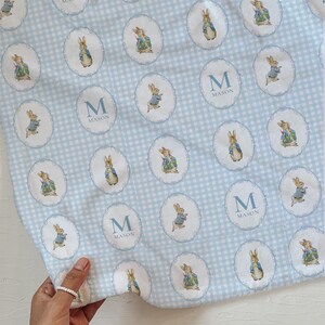 May include: A baby blanket with a light blue and white gingham pattern. The blanket is decorated with oval illustrations of a rabbit in a blue jacket and the name "MASON" with the letter "M".
