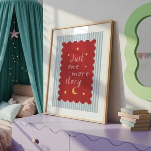 A framed poster with a red background and a blue and white striped border, displaying the text "Just one more story" in a whimsical font. The poster is placed on a purple dresser, with a stack of books and a green mirror visible in the background.