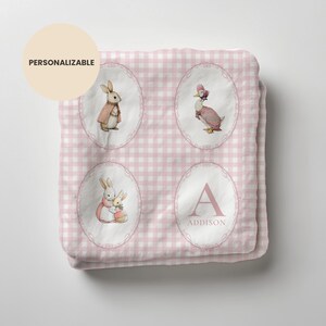 May include: A pink and white gingham baby blanket featuring illustrations of bunnies and a duck in oval frames. One oval has the letter "A" and the name "Addison". The blanket is personalised and the text "PERSONALIZABLE" is in a circle in the upper left corner.