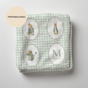 May include: A personalized baby blanket with a light green gingham pattern. The blanket features four oval illustrations of Peter Rabbit and a watering can. The word "PERSONALIZABLE" is in a circle on the top left. The name "MASON" is below the letter "M".