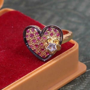 Può includere: Un anello d'oro a forma di cuore con un piccolo design a fiore bianco e pietre rosa. L'anello è in una scatola di velluto rosso.