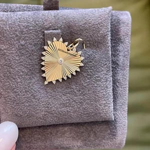 May include: A gold-toned heart-shaped pendant featuring a sunburst pattern with a central clear stone and a border of small clear stones. The text 925SilverEmporium is printed on the surface. It includes a small metal loop for attachment to a chain.