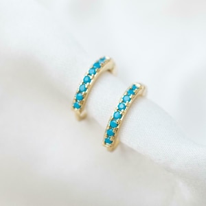May include: A pair of gold-colored hoop earrings, each adorned with a row of small, vibrant turquoise-colored stones. The earrings are set against a soft, white fabric background, highlighting their design and color.
