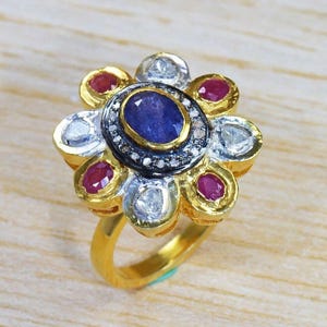May include: A gold ring with a flower design. The centre has a blue oval gemstone, surrounded by diamonds and a black border. Red gemstones and diamonds are set around the centre, with a gold band.