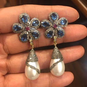 May include: A pair of flower-shaped earrings with blue gemstone petals and a small red gemstone center. The earrings have a pear-shaped pearl drop with a diamond-encrusted cap and a diamond-accented swirl. Jewelry.