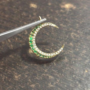 Può includere: Un ciondolo a forma di mezzaluna realizzato in oro, caratterizzato da un design a doppia fila. Una fila è incastonata con piccole pietre preziose rotonde di un verde vibrante, mentre la fila adiacente è tempestata di minuscole pietre bianche.