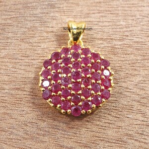 May include: A circular gold pendant featuring a cluster of numerous small, round, deep pink rubies set closely together. The pendant includes a gold bail at the top for attachment. The text 925SilverEmporium is visible above the pendant.