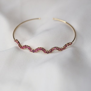 Könnte beinhalten: Ein goldfarbenes Armreif-Armband mit einem wellenförmigen Design. Die Welle ist mit einer Reihe kleiner, leuchtend rosa Edelsteine verziert. Das Armband ist an den Enden offen und liegt auf einem weißen, strukturierten Stoff.