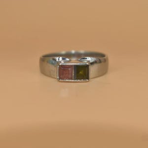May include: A silver ring featuring two square gemstones, one pink and one olive green. The ring has a simple, classic design. The gemstones are set side by side, adding a pop of colour to the piece. A stylish accessory.