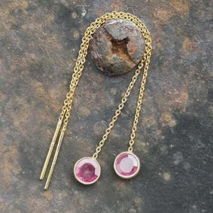 Può includere: Un paio di orecchini a filo dorati con due catenine sottili collegate a perni dritti. Ogni catena termina con una pietra preziosa rosa rotonda incastonata a lunetta. Il design permette alle catene di pendere liberamente con le pietre.