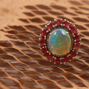 May include: A ring with a large oval opal surrounded by a halo of small red gemstones and diamonds.