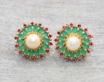 May include: A pair of gold-toned stud earrings. Each earring features a central white pearl surrounded by green and red gemstones, arranged in a floral pattern. The earrings are set against a gray background.