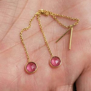 May include: Gold-coloured chain earrings with pink gemstone details. The earrings have a chain that hangs from the ear, with a small, round, pink gemstone at the end of each chain. The earrings are held in a hand.