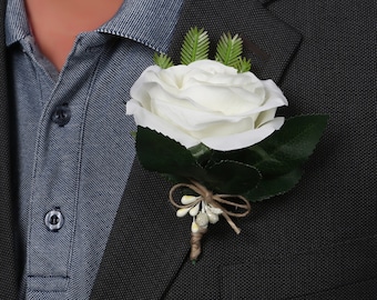 May include: A white rose boutonniere with green leaves and a brown twine bow. The boutonniere is pinned to a dark gray suit jacket.